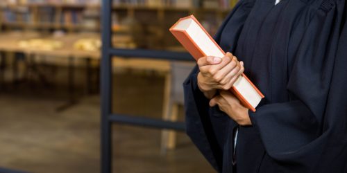 Female student wearing graduation gown standing by glass partition holding orange book in library. Scholar, learning, education, study, academic, research, library